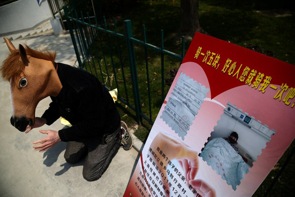 Chen Yuntao, wearing a horse head mask, kneels beside a billboard advertising 5 yuan ($0.8) rides on his back on a road in Hefei, capital city of East Chinas Anhui province April 13, 2015. Chen is trying to raise money to help his 9-year-old son, Chen Minghao, who has leukemia, because his family cannot afford the medical fees, which cost at least 400,000 yuan ($64,440)  a huge sum of money for an ordinary family. Many passers-by donated some money to help the poor father without taking up his offer of a ride on his back.[