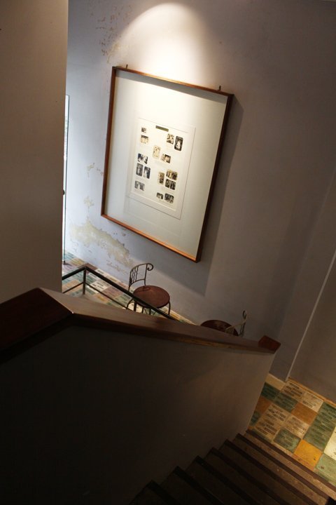 Một góc cầu thang lên tầng lại được đặt trong không gian hơi tối tách biệt hẳn với không gian sáng bên cạnh được treo những bức tranh mang tính hoài niệm.