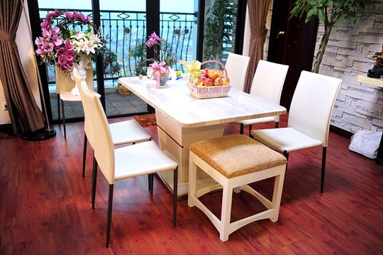 Bộ bàn ăn trắng tinh tế có cùng tông với sofa, nổi bật trên nền sàn gỗ. Nhiều bình hoa lớn, nhỏ được đặt trên bàn và góc phòng khách khiến căn nhà càng thêm rực rỡ
