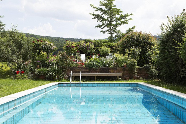 Phía sau nhà là bể bơi rộng và bục bán nguyệt, là nơi trồng thêm những loại hoa rực rỡ. Ở chính giữa được đặt ghế trắng xinh xắn.