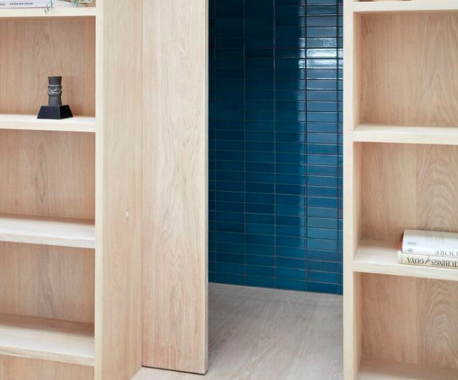 Phía sau kệ đựng đồ nhìn từ khu vực đầu tiên là cánh cửa gỗ sồi màu gỗ sáng trượt sang hai bên, không gian phía trong được bố trí ấn tượng với nền gạch màu xanh lam đậm.