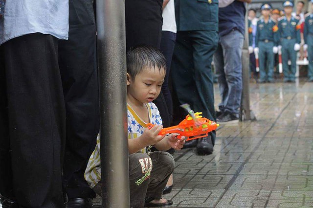 Một bé trai đang nghịch chiếc máy bay mô hình dưới trời mưa như trút nước ngoài sân nhà tang lễ khiến nhiều người xót lòng.