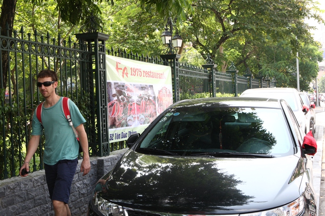 Tấm biển quảng cáo nhà hàng được căng trên tường rào khu di tích Văn Miếu-Quốc tử giám. Xe ô tô đỗ kín trên vỉa hè khiến người đi bộ phải lách.