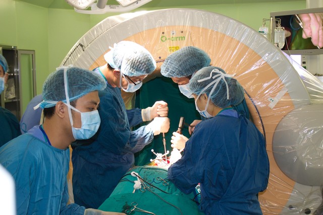 
Các bác sĩ phẫu thuật cột sống cho bệnh nhân bằng hệ thống máy O-arm (màu vàng) tiên tiến. Ảnh: Ami Hà
