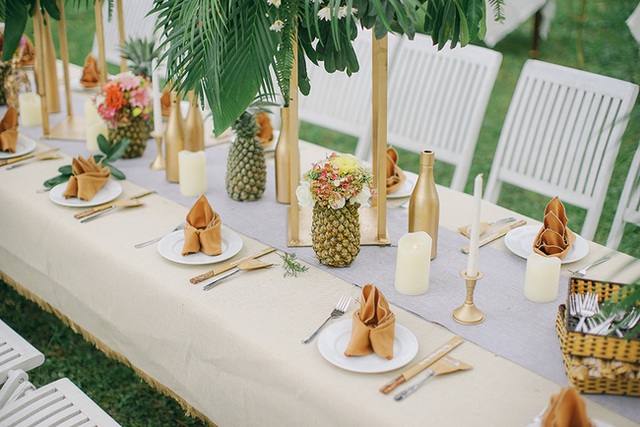 B&agrave;n tiệc được xếp d&agrave;i theo phong c&aacute;ch phương T&acirc;y. Những chiếc lọ thủy tinh được phun sơn m&agrave;u đồng sang trọng, hoa được cắm tr&ecirc;n cao để tr&aacute;nh g&acirc;y vướng v&iacute;u cho kh&aacute;ch khi d&ugrave;ng bữa. Ngo&agrave;i ra, b&agrave;n tiệc c&ograve;n được trang tr&iacute; với những quả thơm v&agrave; những b&igrave;nh hoa từ ch&iacute;nh những quả thơm đ&atilde; được nạo ruột n&agrave;y. V&igrave; tiệc được tổ chức v&agrave;o buổi tối, n&ecirc;n b&agrave;n tiệc c&ograve;n c&oacute; th&ecirc;m nến trắng rất tinh tế v&agrave; đ&aacute;ng y&ecirc;u.