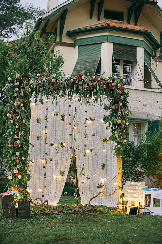 Cổng ch&iacute;nh h&igrave;nh v&ograve;m được tết bằng c&aacute;c loại l&aacute; xanh điểm th&ecirc;m v&agrave;i b&ocirc;ng hoa trắng, khiến cho tiệc cưới c&agrave;ng th&ecirc;m gần gũi với thi&ecirc;n nhi&ecirc;n. Hai b&ecirc;n lối dẫn ch&iacute;nh l&agrave; những giỏ hoa treo xinh xắn bằng quả thơm.