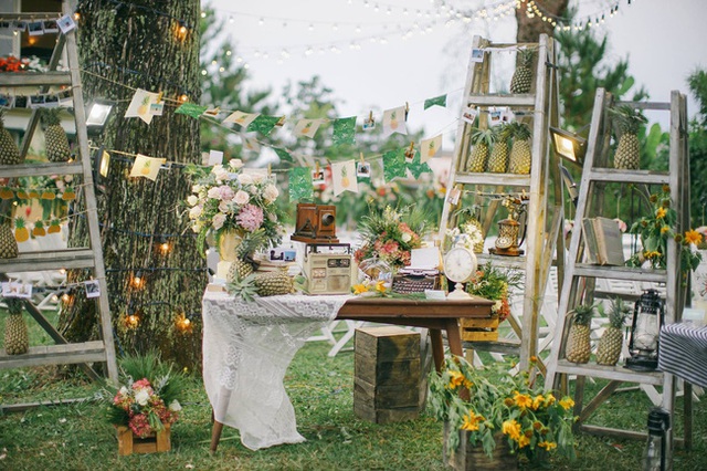 Cờ giấy in h&igrave;nh quả thơm xen l&atilde;nh m&agrave;u xanh nhẹ nh&agrave;ng đậm chất vintage. M&aacute;y ảnh, đ&agrave;i cassette, điện thoại, đồng hồ kiểu cổ v&agrave; v&agrave;i cuốn s&aacute;ch cũ đ&atilde; tạo n&ecirc;n một chiếc b&agrave;n trang tr&iacute; độc đ&aacute;o ngay lối v&agrave;o. Những chiếc thang gỗ v&agrave; thang sắt mộc mạc cũng được sử dụng để b&agrave;y hoa d&atilde; quỳ, s&aacute;ch v&agrave; những quả thơm trang tr&iacute;.