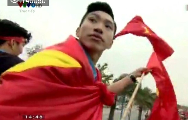 Văn Hậu khoác cờ Việt Nam nhưng tay không quên cầm một lá cờ khác vẫy chào cổ động viên. Ảnh cắt từ clip.