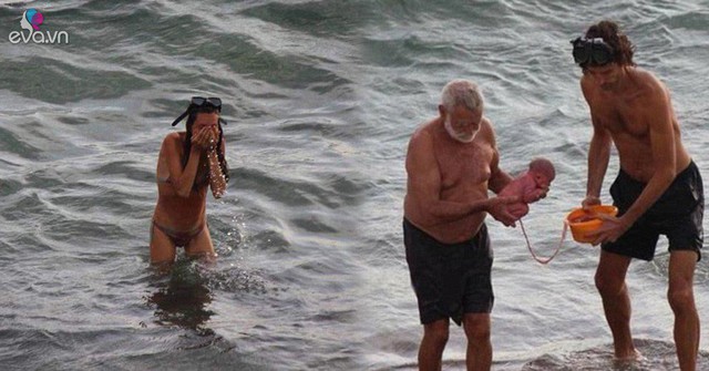 Hai người đàn ông ôm một bé sơ sinh còn nối nguyên với nhau thai đi từ biển vào bờ.