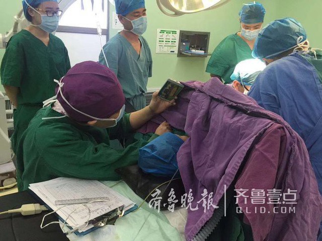 Y t&aacute; Trương Tuệ quỳ b&ecirc;n b&agrave;n mổ để cầm điện thoại cho b&eacute; trai xem hoạt h&igrave;nh. (Ảnh: Qlid)