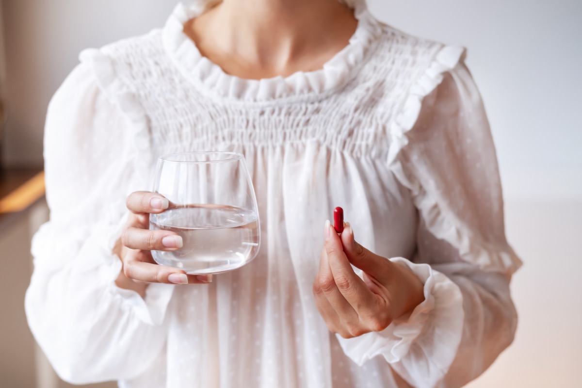 Người phụ nữ mặc áo trắng cầm một viên thuốc màu đỏ trong một tay và tay kia cầm ly nước